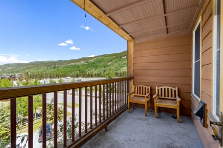 A covered balcony with wooden chairs overlooks a parking area and a landscape of green hills under a clear blue sky.