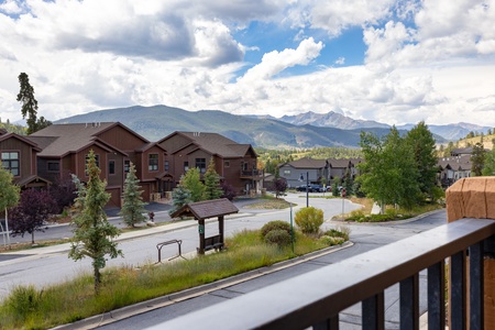 A scenic view of a suburban neighborhood with brown wooden houses, a mountainous backdrop, and a cloudy sky. Trees and a street sign are visible in the foreground.