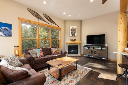 A cozy living room with leather sofas, a wooden coffee table, a flat-screen TV, a fireplace, large windows, and wall-mounted snowshoes. The room is decorated with nature-themed art and a patterned rug.