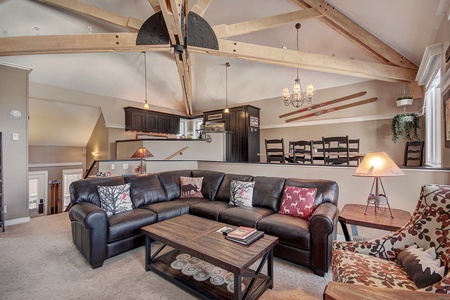 A cozy living room featuring a large black leather sectional, a wooden coffee table, and a patterned armchair. The room has exposed wooden beams, a chandelier, and decorative skis on the wall.