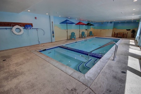 Indoor pool area with a small rectangular pool, beach mural on the wall, four colorful umbrellas, lounge chairs, and poolside safety equipment. Natural light is coming in through windows on the right.