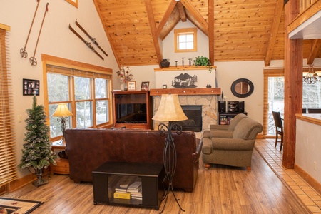 Cozy living room with wooden beam ceiling, fireplace, and large windows. Brown leather sofa, armchair, and TV stand are arranged around the fireplace. Ski décor and lamps add to the warm ambiance.