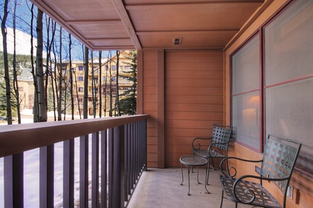 Outdoor balcony with two metal chairs and a small table. Wooden railing and siding visible. Snow-covered ground and buildings in the background. Trees without leaves lining the perimeter.