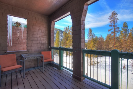 A covered balcony with two orange chairs, a small table, icicles hanging from the roof, and a snowy forest landscape visible through the wooden railing and arches.