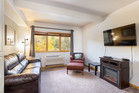 A cozy living room with a leather sofa, chaise lounge, and wall-mounted TV. The room features a large window with a view of autumn foliage, and a fireplace beneath the TV adds warmth.