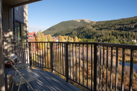 A wooden balcony with two chairs overlooks a scenic view of trees, hills, and buildings under a clear blue sky.