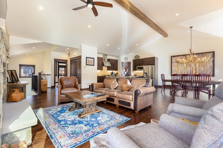 Spacious living room with vaulted ceiling, wooden beams, and a ceiling fan. Features leather sofas, armchairs, a coffee table, a fireplace, and an adjacent dining area with a large table and chandelier.