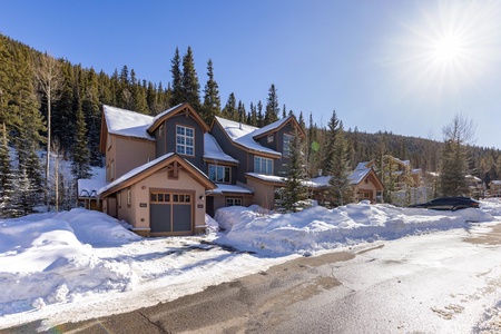 A snow-covered street in a mountain village features houses with sloped roofs and snow piled on the sides. Tall evergreen trees stand in the background under a clear blue sky with the sun shining.