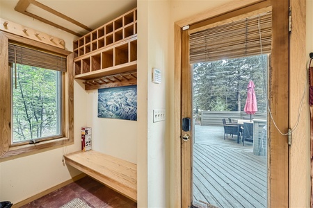 A mudroom with wooden shelves, a bench, and a window on the left, and a glass door leading to an outdoor deck with a table and umbrella on the right.