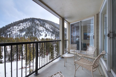 A balcony with metal chairs and a table overlooking a snow-covered mountain landscape with pine trees.