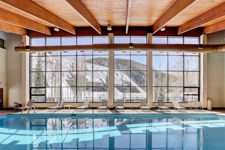 Indoor pool area with large windows showcasing a snowy mountain landscape. The space includes lounge chairs and wooden ceiling beams.