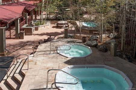 Outdoor area with two hot tubs, a patio, and surrounding trees. The setting features stone decking, metal railings, and a nearby red-roofed building.