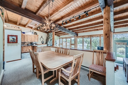 A dining room with a wooden table and chairs, an antler chandelier, and wooden beams on the ceiling. The room opens to a kitchen and features large windows with a view of the outdoors.