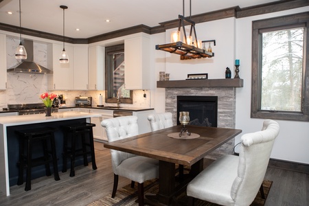 A modern kitchen and dining area with white cabinets, marble backsplash, dining table with four upholstered chairs, black stove hood, island with stools, and a stone fireplace with a candle holder.