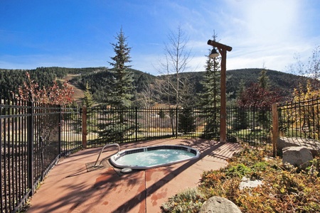 Hot tub on a concrete patio, surrounded by a black metal fence. The backdrop consists of a forest and mountains under a clear blue sky.