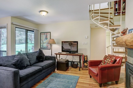 A cozy living room features a gray sofa, red armchair, wooden coffee table, wall-mounted TV, spiral staircase, and large window with a scenic view. A framed poster decorates the wall.