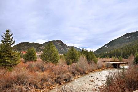 A serene landscape featuring a mountain range, a river flowing through a valley, and a small bridge crossing the river amidst trees and shrubs.