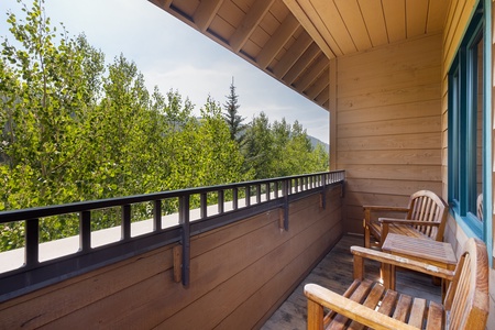 A wooden deck with chairs overlooks green trees and a forested landscape. The deck has a wooden ceiling and railing.