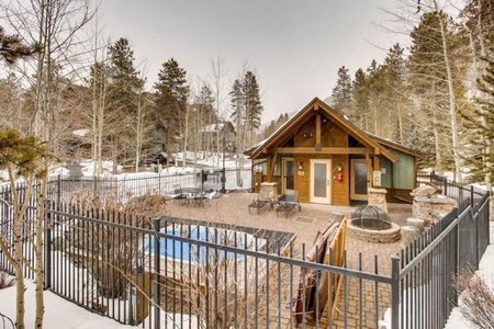 A small fenced-in area with a jacuzzi and outdoor seating in front of a rustic cabin surrounded by snow-covered trees.