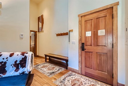 Entryway with a wooden door and a bench against the wall. There are decorative rugs on the floor and a coat rack with hooks above the bench. A partially visible piece of furniture has a cowhide throw.