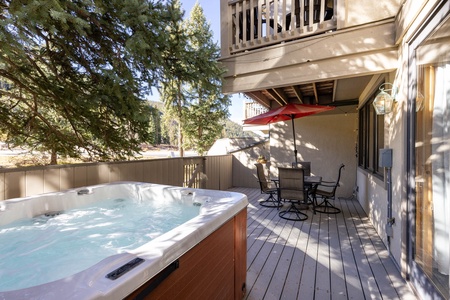 Deck with a hot tub and a patio set under a red umbrella, surrounded by trees and shaded by an above balcony.