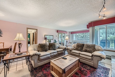 A living room with two beige sofas, fur pillows, a wooden coffee table, a lamp, a large window with forest views, and a red-patterned rug. Wall decor and a small table with chairs are visible.