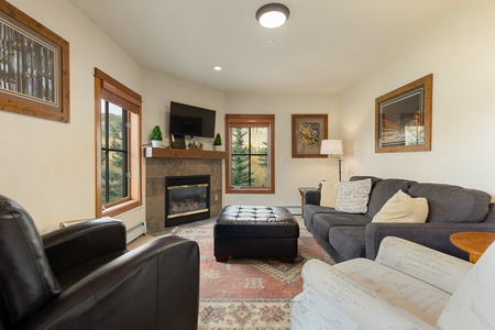 A cozy living room with a gray sofa, black leather armchair, fireplace, wall-mounted TV, and large windows. Decor includes framed art, a floor lamp, and a patterned area rug.