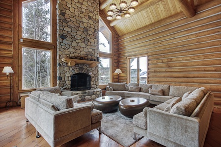 Spacious living room featuring a stone fireplace, large sectional sofas, wooden ceiling with beams, and floor-to-ceiling windows with views of the trees outside.