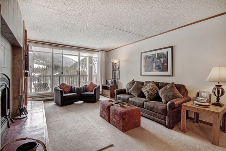 A cozy living room with a brown sofa, two armchairs, a coffee table, and a fireplace. Large windows offer a view of a snowy landscape with mountains. The room is decorated with a framed picture and a lamp.