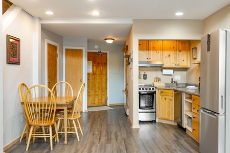 A small kitchen with wooden cabinets, a stainless steel refrigerator, a stove, and a sink. Adjacent is a dining area with a wooden table and four chairs. Vinyl flooring runs throughout the space.