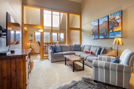 A cozy living room with a large grey sectional sofa, a striped armchair, and a wooden coffee table. Large windows illuminate the space. A painting of a buffalo is on the wall.