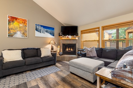 A cozy living room features a fireplace, two gray sofas, and a large ottoman. Art pieces and a TV are mounted above the fireplace. Windows on the right wall provide natural light and an outdoor view.