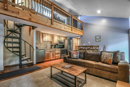 Living area with a sofa, coffee table, open kitchen with wooden cabinets, and a spiral staircase leading to a loft. The loft features a wooden railing. Natural light fills the space.