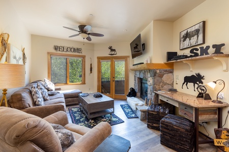 A cozy living room with beige furniture, a fireplace, and a TV mounted above it. Decorative accents include moose-themed items and a "Welcome" sign. Large windows and a door offer natural light.