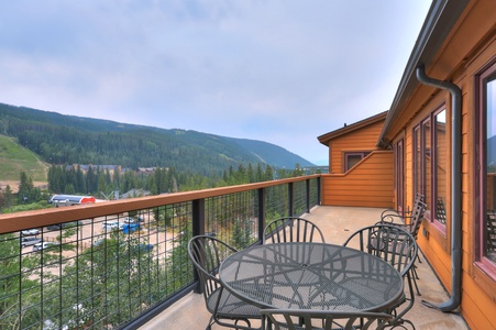 A balcony with a metal table and chairs overlooks a mountainous landscape, pine trees, and buildings below on a clear day.