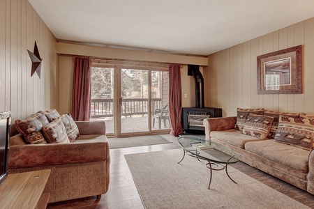 A living room with wood-paneled walls, two sofas with patterned cushions, a glass coffee table, a TV, a framed picture, and a wood stove next to large sliding glass doors leading to a deck.