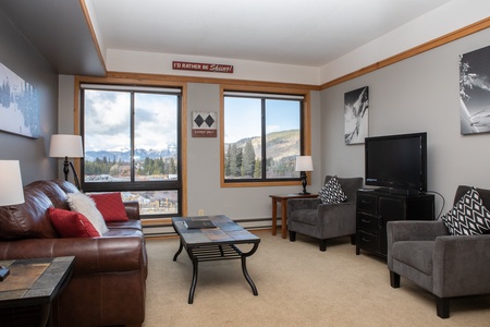 A cozy living room with a leather sofa, two armchairs, a coffee table, and a TV. Large windows offer a view of mountains and trees. Black-and-white artwork decorates the grey walls.