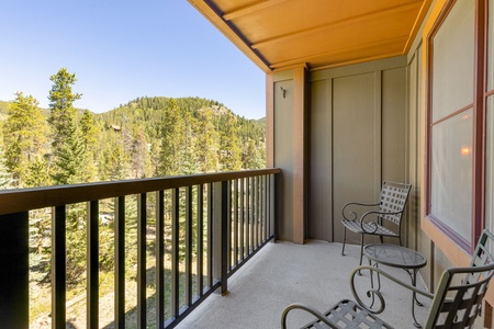 Balcony with two metal chairs and a small table overlooking a scenic view of a forested mountain landscape on a clear day.