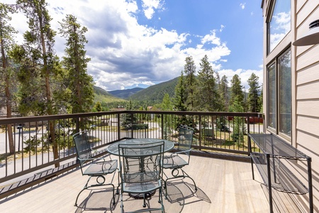 A wooden deck with a metal table and chairs overlooks a forested landscape with mountains in the background under a partly cloudy sky.