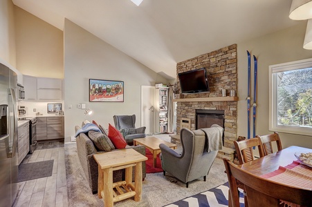 A cozy living room with a stone fireplace, TV above it, and a mix of modern and rustic furniture. The kitchen is visible on the left, and ski equipment is propped against the wall near a large window.