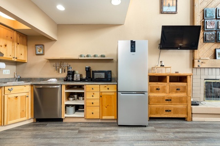 A modern kitchen with wooden cabinets, stainless steel appliances including a refrigerator and dishwasher, a microwave, coffee maker, and a wall-mounted TV beside a fireplace.