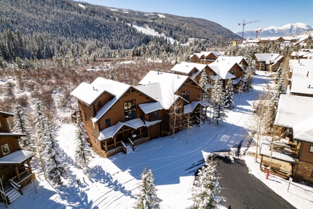 A snowy hillside community with wooden houses, surrounded by pine trees. A mountain range is visible in the background under a clear, blue sky.