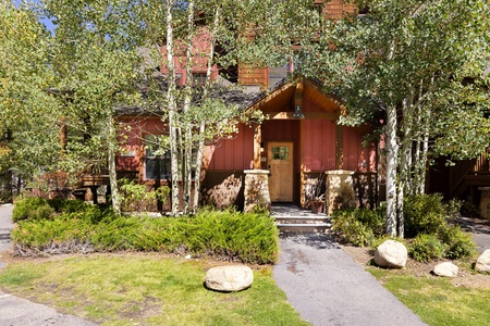 A red wooden house with a stone foundation and covered porch, surrounded by trees and shrubs, viewed from the front. The house is located in a wooded area with a concrete path leading to the entrance.