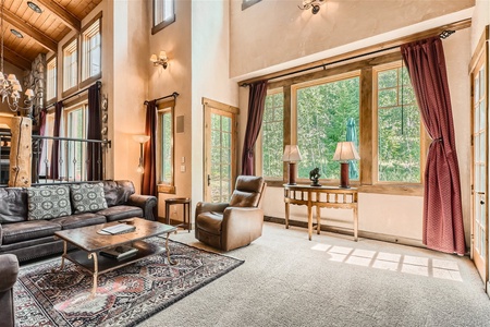 A well-lit living room with a high ceiling, large windows, a dark leather couch, two armchairs, and a wooden coffee table. The space has light walls, a patterned rug, and draped curtains.