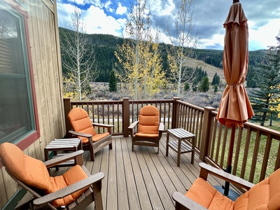 Wooden deck with four orange cushioned chairs, two side tables, and a closed umbrella. Overlooks a forested mountain landscape under a partly cloudy sky.