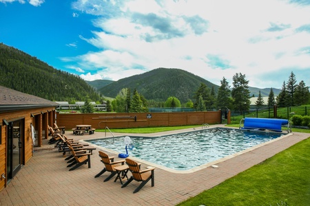 A tranquil outdoor swimming pool surrounded by wooden lounge chairs and a wooden fence, with green hills and a blue sky with clouds in the background.