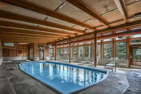 Indoor swimming pool area with wooden ceiling beams, large windows, lounge chairs, and a jacuzzi in the corner.