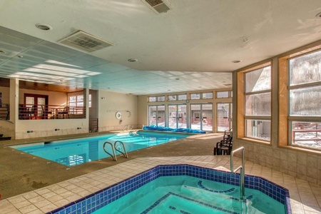 Indoor swimming pool area with an adjacent hot tub, surrounded by large windows allowing natural light. The space includes seating and handrails for safety.