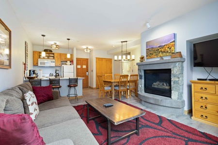 A cozy living room with a stone fireplace, a wall-mounted TV, a gray sectional sofa, and a wooden coffee table. The open kitchen features wooden cabinets and a dining area with a table and chairs.