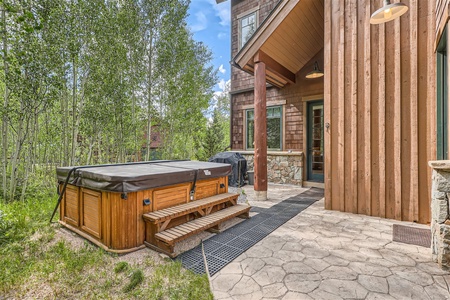 Outdoor area with a wooden hot tub, stone paver patio, and steps leading to a wooden house. Surrounded by trees and greenery in a natural setting.